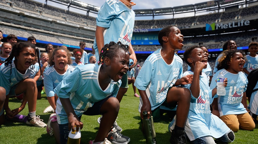 a group of youth have their mouths open as they scream in excitement. there are four black boys at the front who are sitting or kneeling on their knees. there are a couple of girls in view just behind them. all of them are black and are wearing light blue U.S. Soccer Foundation jerseys.